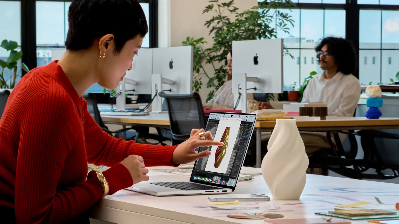 Žena v červeném svetru sedí u stolu v kanceláři a prstem se dotýká displeje MacBooku Pro, na kterém je 3D model vázy, stejné, jaká stojí vedle na stole