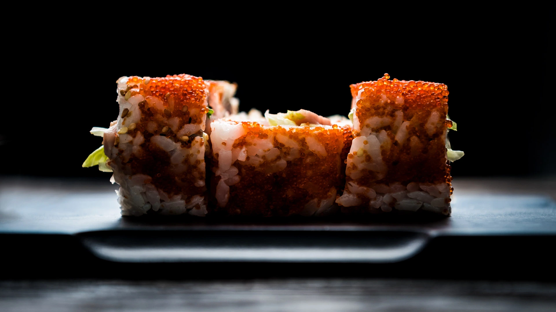 Nasvícené sushi rolky s lososovými jikrami naaranžované na tmavém keramickém talíři